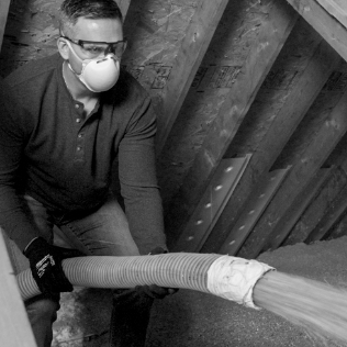 woman with a mask and protective eye gear blowing cellulose insulation in an attic through a hose